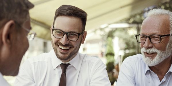 Three men in white shirts and glasses engaged in a friendly conversation outdoors.