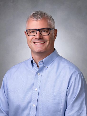 Smiling man in glasses and light blue shirt against a gray background.