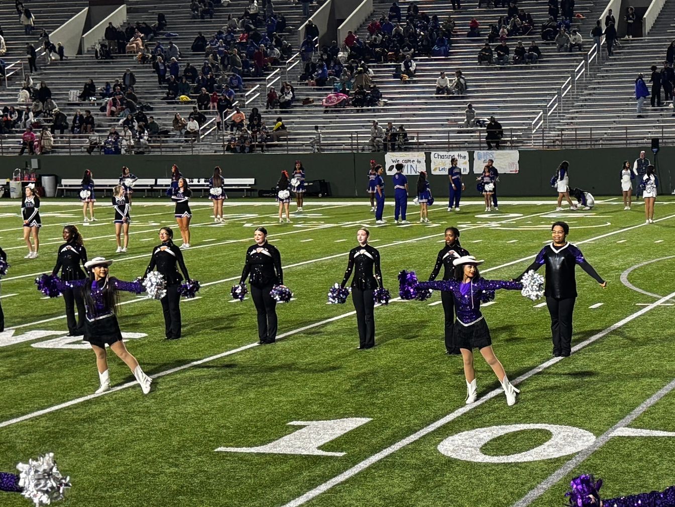 Cheerleaders and dancers perform on a football field with spectators in the stands.