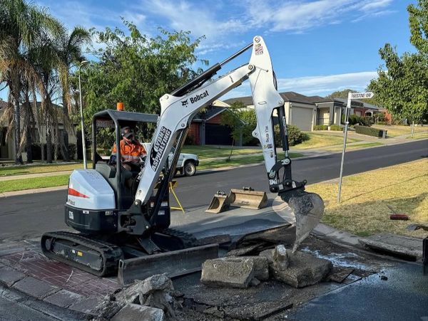 A small excavator breaking concrete on a residential street corner.