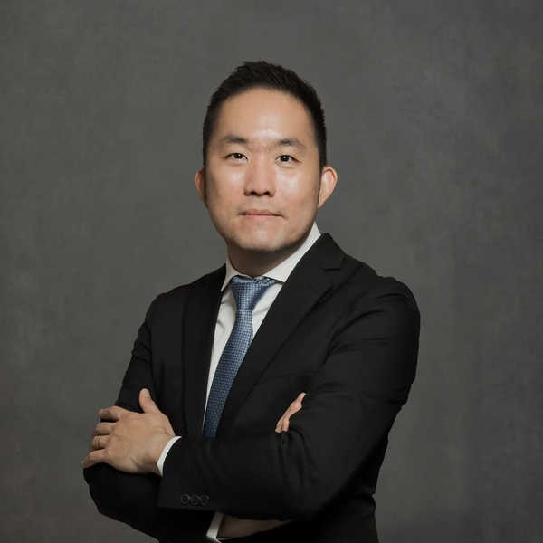 Confident man in a black suit with arms crossed against a grey background.