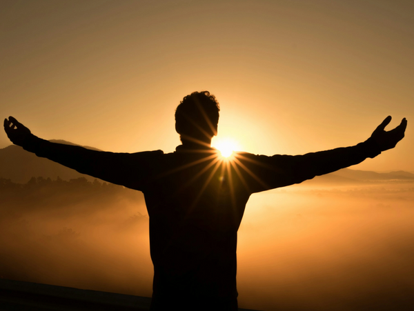 Silhouette of a person with arms wide open facing sunrise over misty hills.