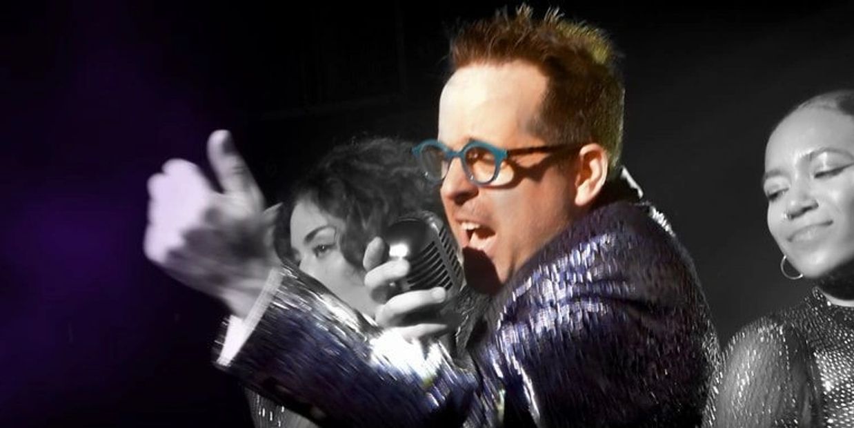 A man passionately singing into a microphone with two backup singers.