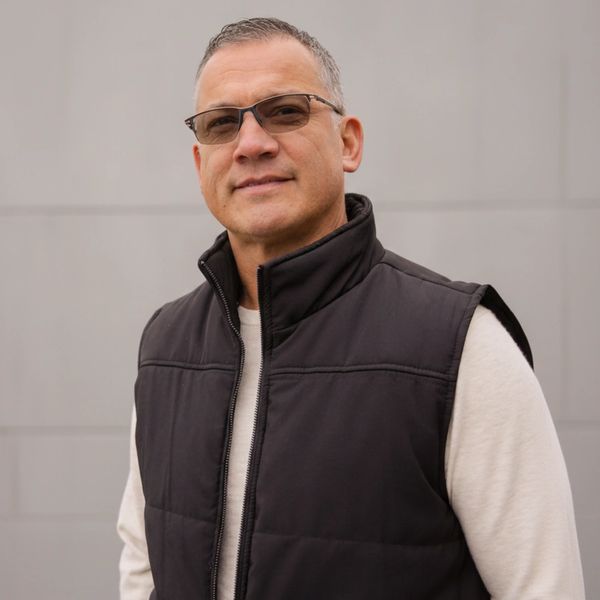 Man wearing glasses and a black vest standing against a gray wall.