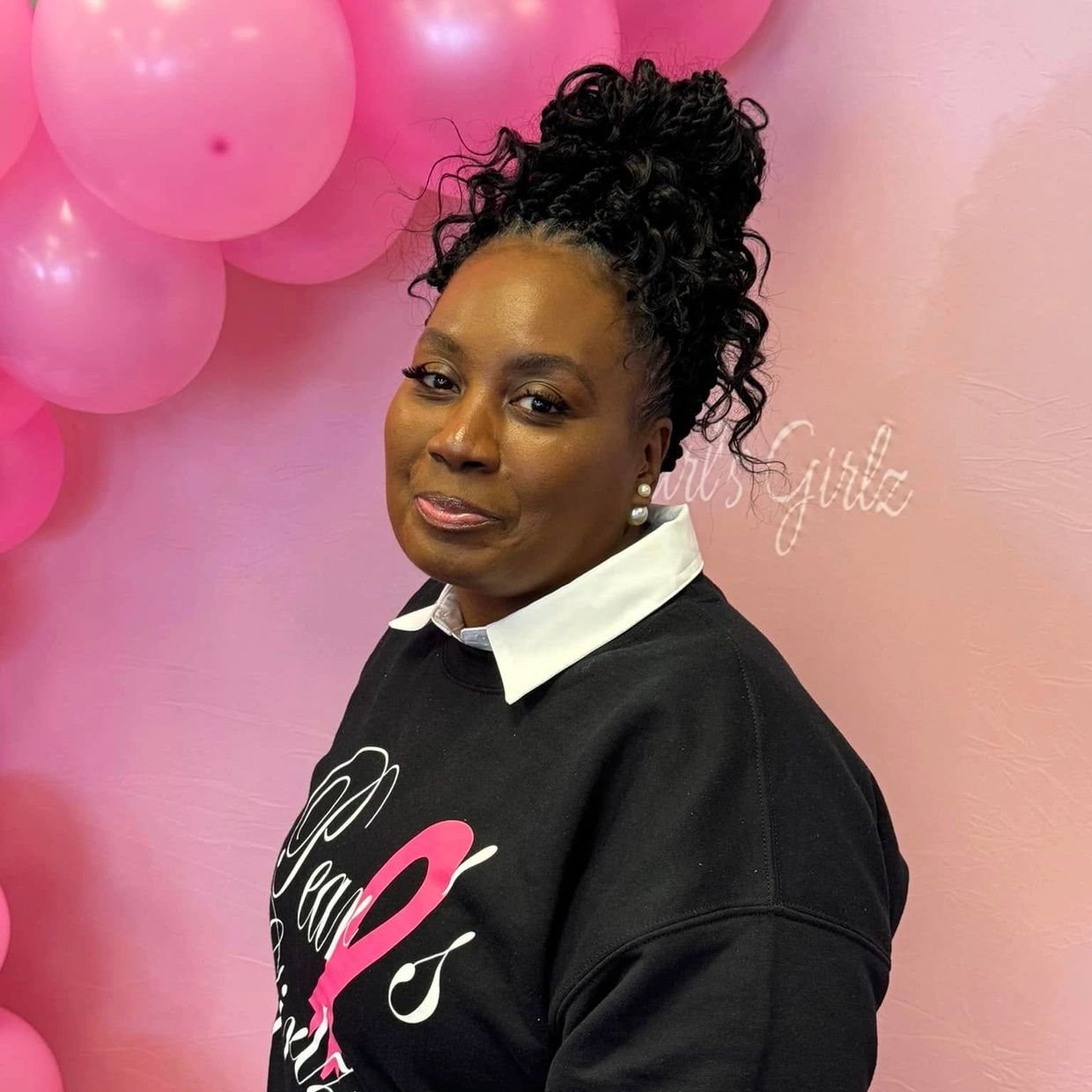 Woman with curly updo hairstyle poses against a pink balloon backdrop.