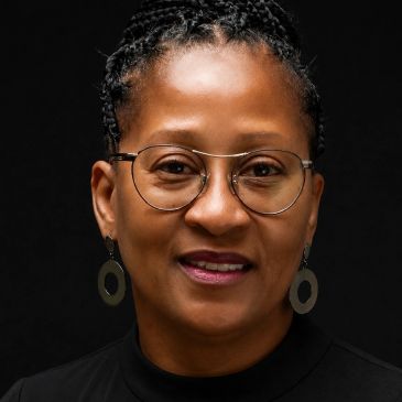 Portrait of a confident woman with glasses and hoop earrings against a black background.