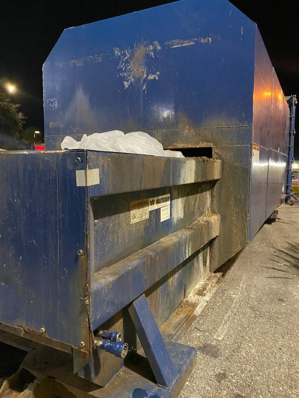 Large blue industrial waste compactor with plastic bags inside, photographed at night.