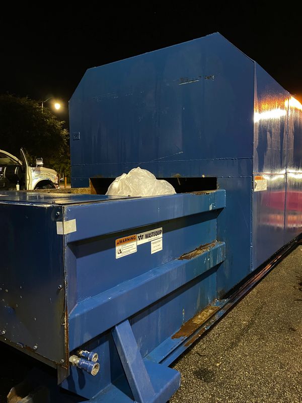 Large blue waste compactor with a plastic bag inside, photographed at night.