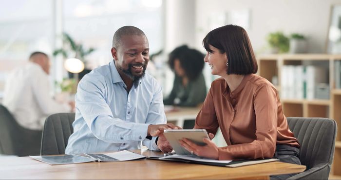 Two colleagues happily discussing work using a tablet in a modern office.