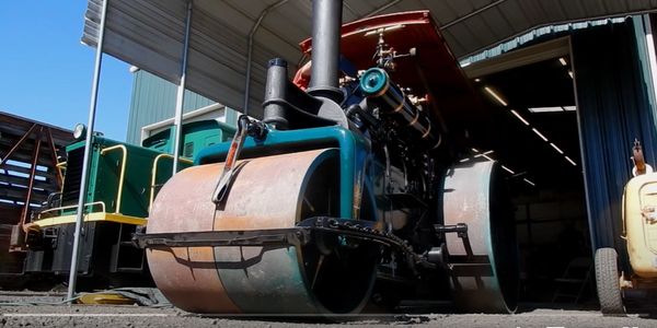 Vintage steamroller parked under a metal roof in a workshop.