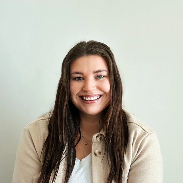 Smiling woman with long brown hair wearing a beige jacket and white top.