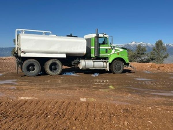 Green and white water tanker truck on muddy construction site.