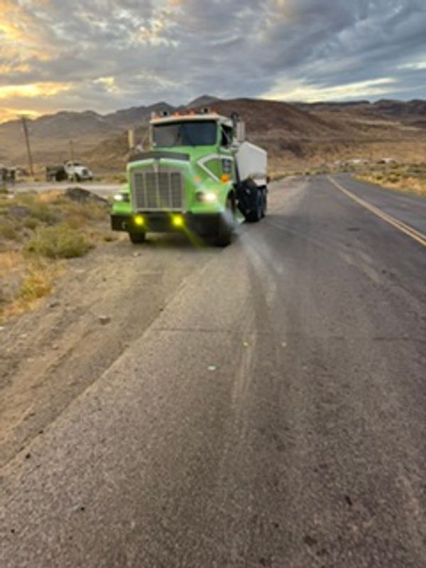 Green street sweeper truck cleaning a desert road at sunset.
