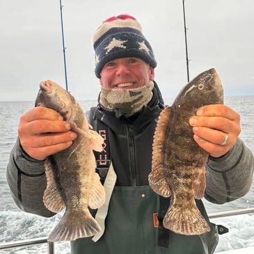 Smiling fisherman holding two large fish on a boat in cold weather.