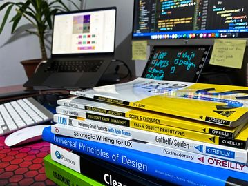 Stack of programming and design books on a desk with coding on screens.