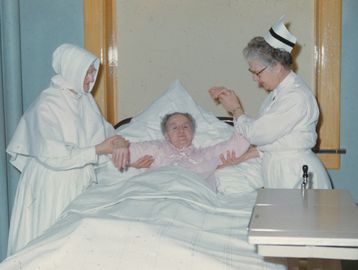 Two nurses assist an elderly woman in a hospital bed.