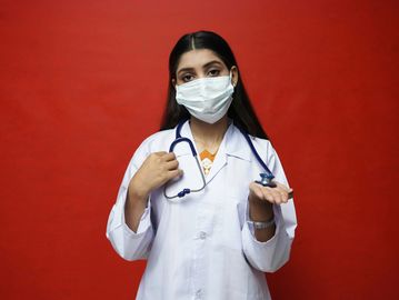 Female doctor in white coat and mask holding a stethoscope against a red background.