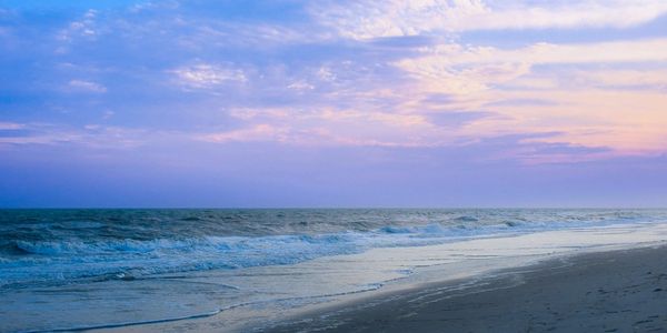 Calm ocean waves gently wash onto a sandy beach under a pastel sunset sky.