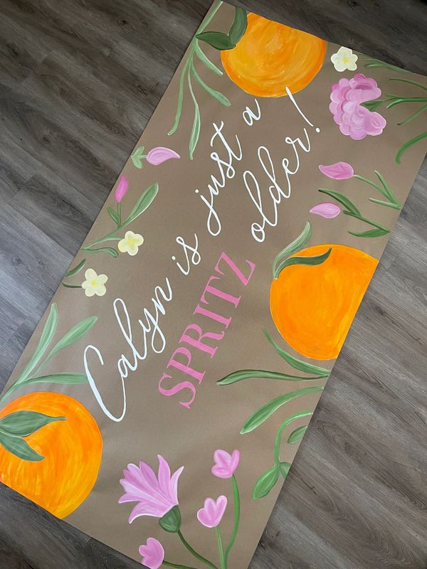 Decorative birthday banner with floral and orange designs saying "Calyn is just a SPRITZ older!"