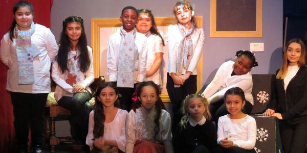 A group of children and a young woman posing indoors, dressed in white tops and scarves.