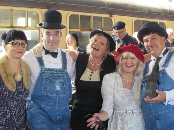 Group of people dressed in vintage and quirky outfits, smiling at a train station.