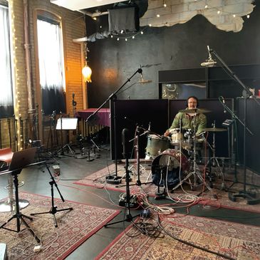 A drummer recording in a cozy, carpeted studio with multiple microphones and instruments.