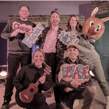 Group of five smiling people holding colorful word signs and a ukulele indoors.