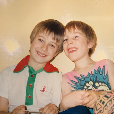 Two young boys smiling and sitting closely against a plain yellow wall.