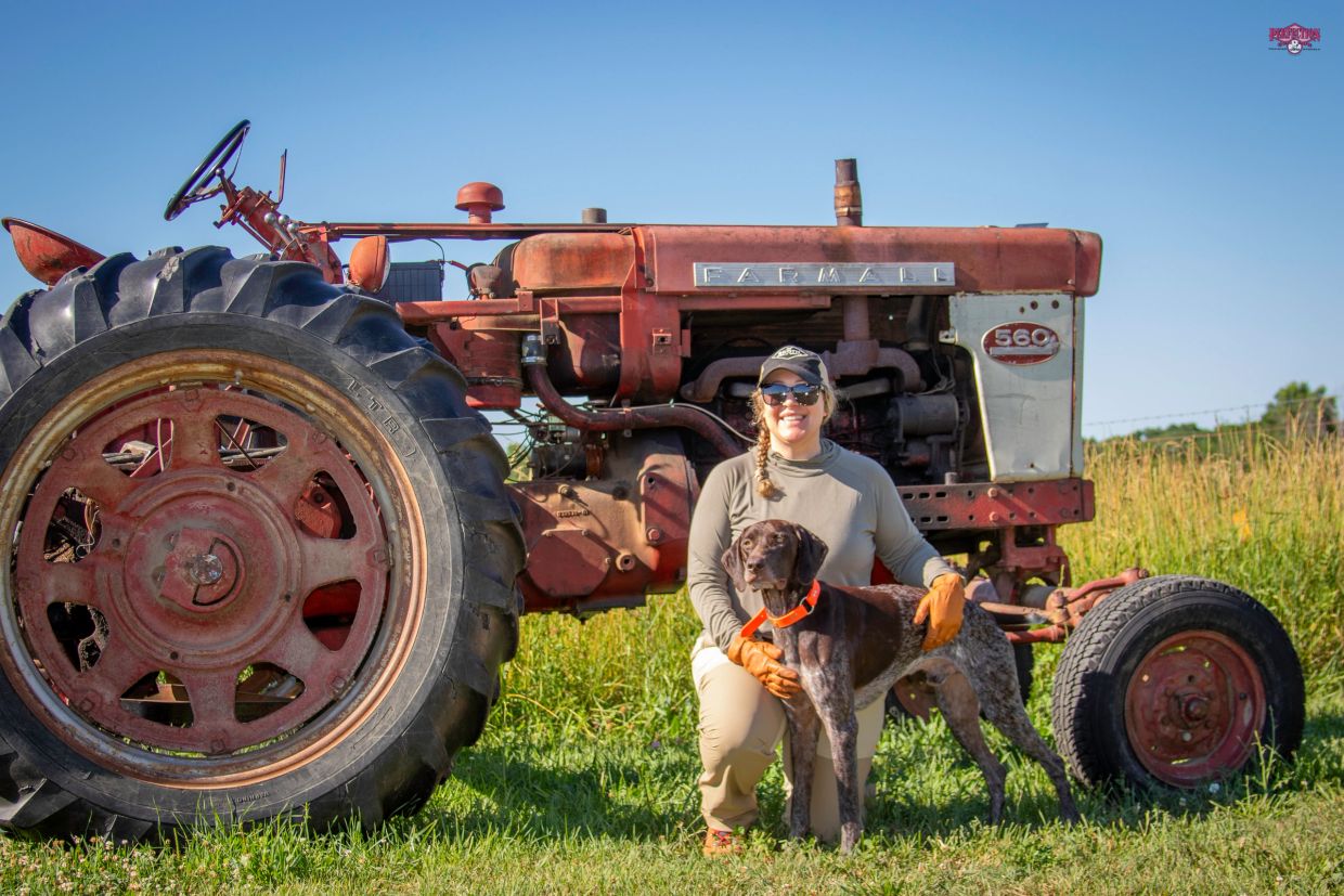 A woman with a dog poses in front of a vintage Farmall tractor on a sunny day.