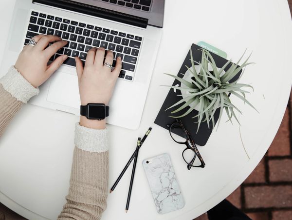 Woman wearing an Apple watch typing on a computer with a book, glasses, phone, pencils and a plant.