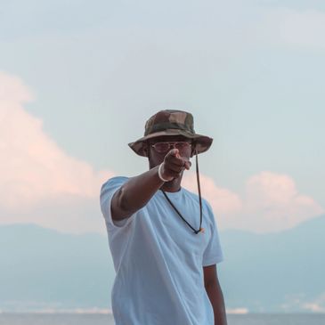 Man pointing at you (the camera) by the beach with a ship in the background.