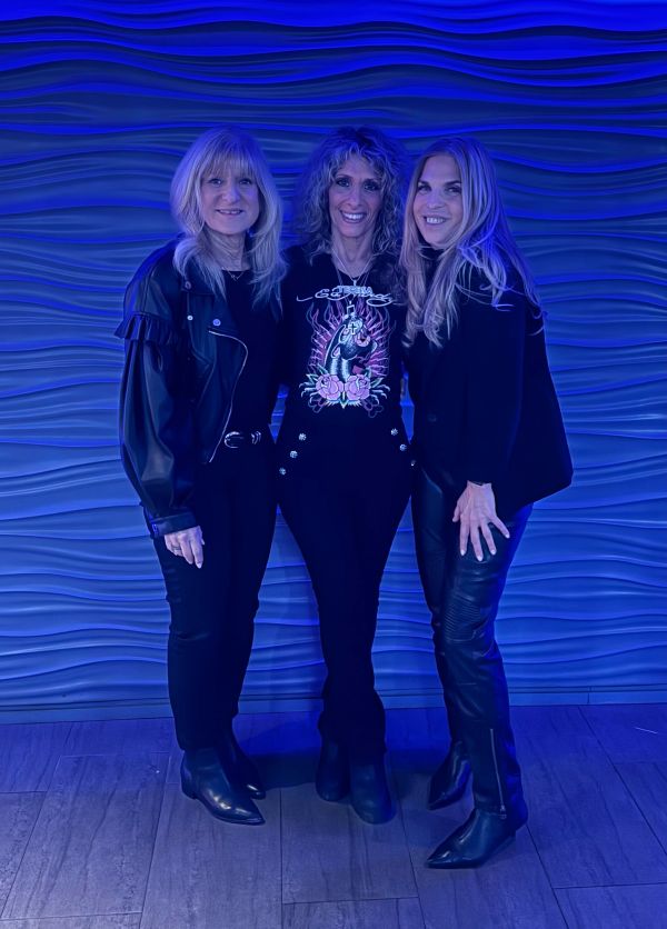 Three women dressed in black with a blue-lit wavy background.