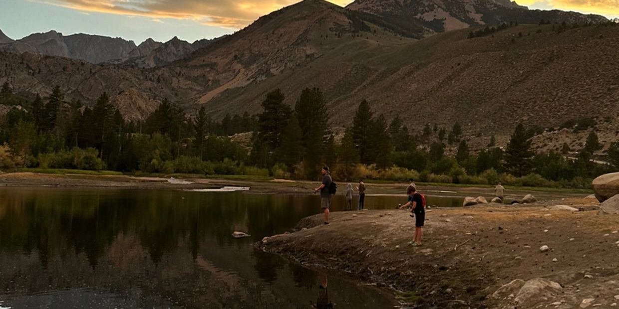 People fishing by a lake with mountains in the background at sunset.