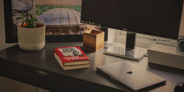 A tidy desk with a laptop, book, plant, and monitor near a window.