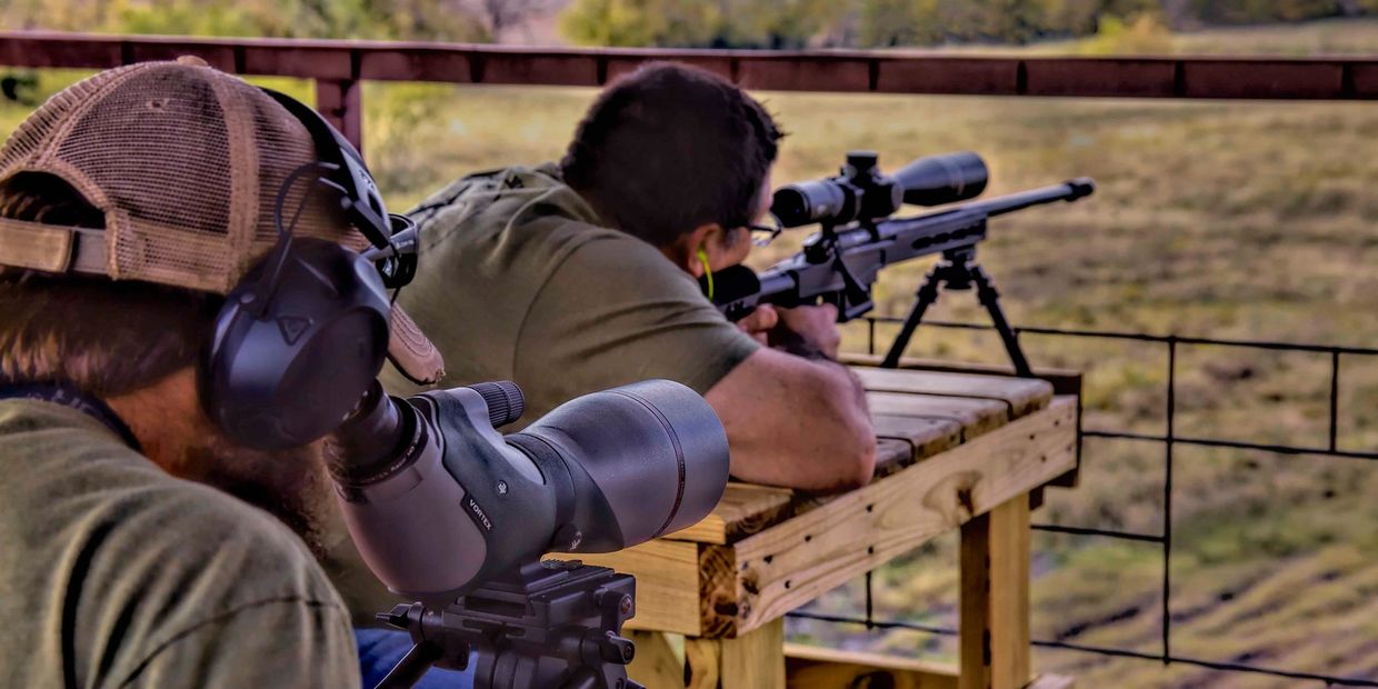 guy siting at a bench shooting a rifle while another guy spots for him