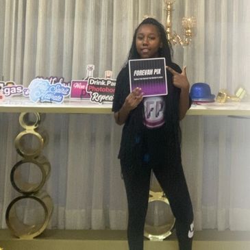 Young woman holding a 'FOREVAH PIX' sign in front of a photo booth setup.