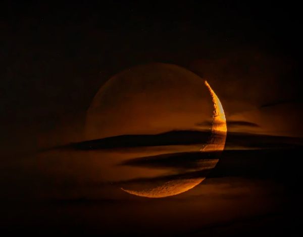 A crescent moon glowing orange behind dark clouds at night.