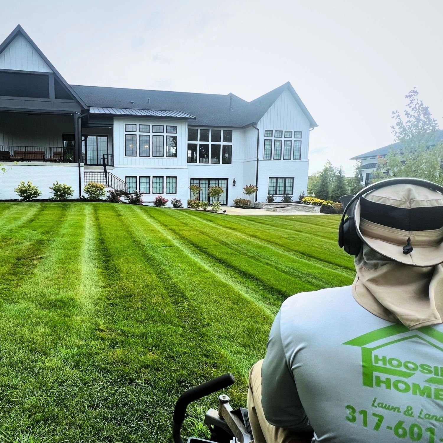 Person mowing a lush, striped lawn in front of a large modern house.