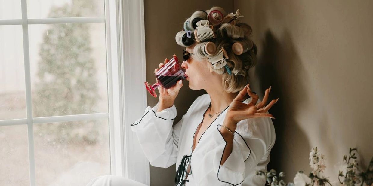 Woman with hair rollers sipping wine by a window.