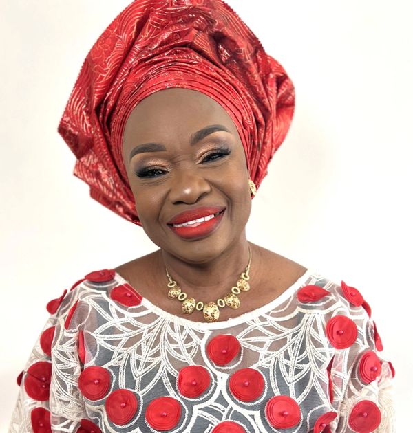 Smiling woman wearing a red traditional headwrap and white dress with red floral details.