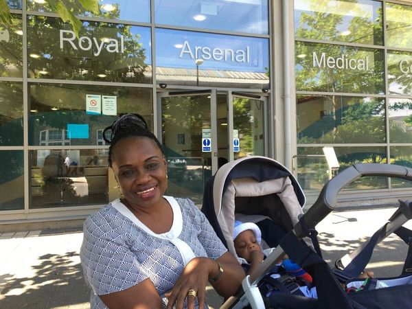 A smiling woman with a baby in a stroller outside Royal Arsenal Medical Centre.