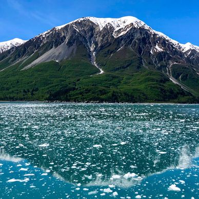 An Alaskan mountain rises into a pale blue sky and is reflected in the glacial icy waters below