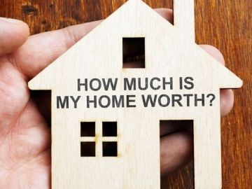 A hand holding a wooden miniature house where the words "HOW MUCH IS MY HOME WORTH?" was written
