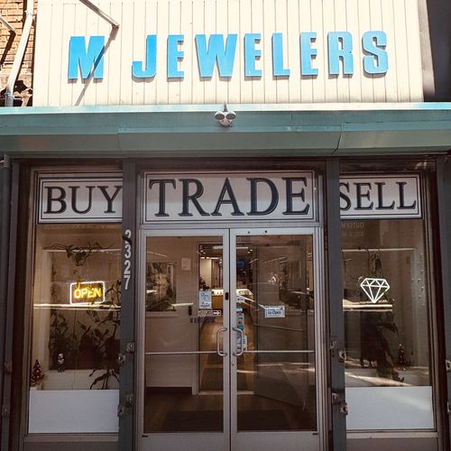Front entrance of M Jewelers with signs for buy, trade, sell, and an open neon sign.