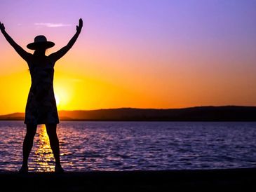 Silhouette of a person with raised arms by the water at sunset.
