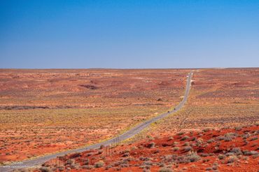 A long road stretches through a vast red desert landscape under a clear blue sky.