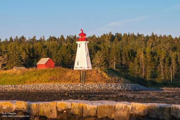 A lighthouse and red building by a forest under a clear blue sky.