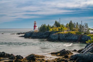 Lighthouse on a rocky coastline with a forest backdrop and calm sea under a partly cloudy sky.