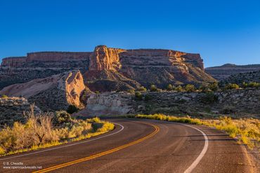Curving road leading to sunlit rocky cliffs under a clear blue sky.