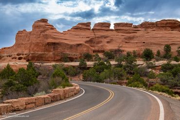 Winding road through red rock formations and greenery under a cloudy sky.